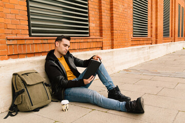 Young student man sitting on a city street reading a book outdoors with a coffee. reading and lifestyle concept.