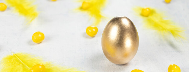 banner with golden egg on a white concrete background with yellow feathers and yellow chocolates in the shape of an egg. Happy Easter concept.