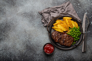 Grilled meat beef steak with potato chips french fries and green peas served with red tomato sauce on rustic metal plate on stone background table from above with space for text