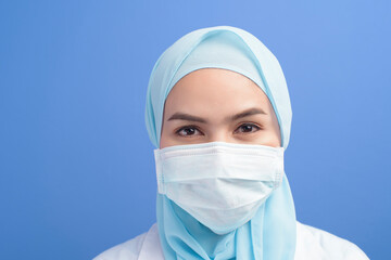 Female muslim doctor with hijab wearing a surgical mask over blue background studio.