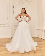 Fototapeta premium Elegant caucasian woman with natural make-up and hairstyle wearing white bridel dress, standing and posing in bright stylish interior. Full length portrait