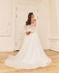Fototapeta premium Elegant caucasian woman with natural make-up and hairstyle wearing white bridel dress, standing and posing in bright stylish interior. Full length portrait