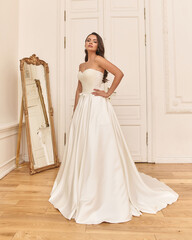 Fototapeta premium Young elegant bride in white luxury wedding dress standing and posing in bright stylish interior with old mirror