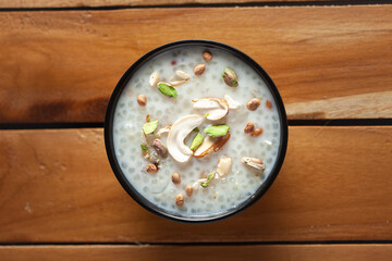 Close-up of Creamy Sabudana Kheer Garnished with dry fruits. Indian delicious dessert. Served in -black ceramic bowl. Top View on wooden background.