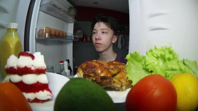 A teenager searches the refrigerator for food. Makes a choice: smoked chicken or a piece of cake .
