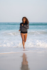 Woman with dark skin and long curly hair having fun at the beach running playing in the waves