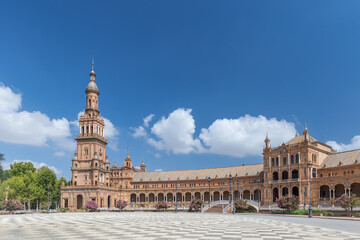 Fototapeta premium Spanish Square, in the center of Seville. Very touristic travel destination empty due to coronavirus measures. View of the south tower. Coloured flowers, blue sky with few clouds. 