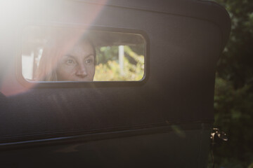 Sad woman looking through rear window of classic car. Nostalgic middle aged lady with thoughtful look. Sun flare, missing past, old times concepts