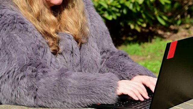 Frau in einer Kaninchenfell Jacke arbeitet im Garten mit einem Notebook im Homeoffice  