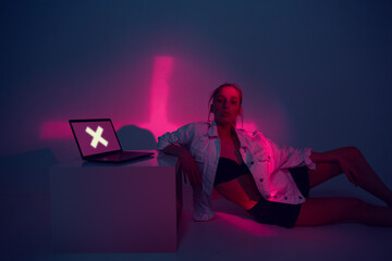 Cool girl with laptop. Cross on screen. Young blogger in home office. Colored Neon light