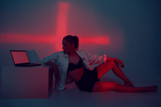 Creative Cool Young Girl With Laptop In Red Studio Light Sitting On The Floor. Fashion Model Blogger With Computer.