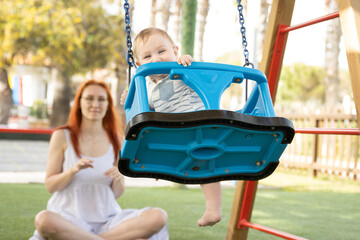 A little funny baby on swings on an outside playground with his ginger mother