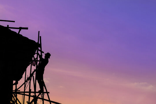 Silhouette construction worker without safety line or harness climbing on D.I.Y. bamboo scaffolding and repair the house roof with dusk sky background. Image for hard working or Labor Day concept.