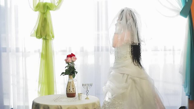 A Jewish bride in a wedding dress and a veil wearing a medical mask in a synagogue hall before the chupah ceremony happily speaks on the phone. Overall plan