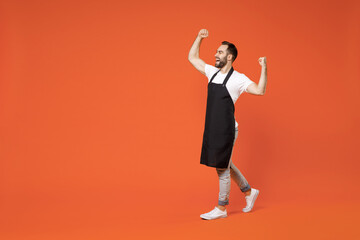Fototapeta na wymiar Full length young man 20s barista bartender barman employee in black apron white t-shirt work in coffee shop do winner gesture clench fist isolated on orange background studio Small business startup.