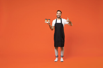 Fototapeta na wymiar Full length young man barista barman employee in apron white t-shirt work in coffee shop hold paper takeaway delivery cups show thumb up gesture isolated on orange background Small business startup.