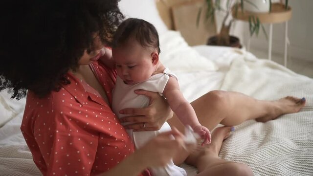 Woman giving pacifier to little baby and hugging him, sitting on bed in apartment room spbd.