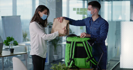 Obraz premium Young caucasian courier in respiratory mask visiting corporate office workspace, delivering food package to young beautiful woman. Lockdown. Services.