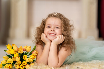 Obraz na płótnie Canvas Portrait cute little girl in beautiful mint-colored dress with wavy hair and a tiara. baby is smiling happily and enjoying the flowers in the background of the fireplace, with a bouquet of tulips.