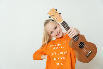 Teenage, blond girl in blue T-shirt is holding light brown ukulele instrument
