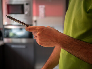 Spanish man dressed in green sending a message with his mobile phone in the kitchen of his house