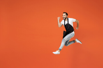 Fototapeta na wymiar Full length side view young fun man barista bartender barman employee in black apron white t-shirt work in coffee shop jump high run fast isolated on orange background. Small business startup concept.