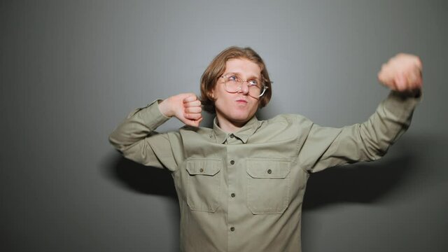 Happy young man in a shirt against a gray background, rejoices in victory with raised arms and dances with a funny face