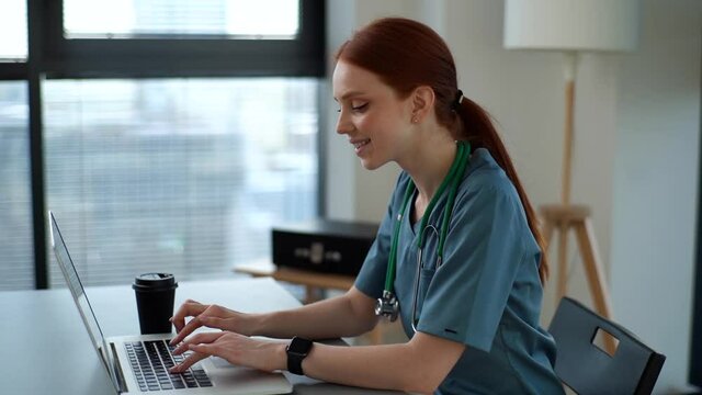 Cheerful Smiling Young Female Doctor In Blue Green Medical Uniform Typing On Laptop Computer Looking On Screen Sitting At Desk Near Window In Modern Office Of Medic Clinic. Shooting In Slow Motion.