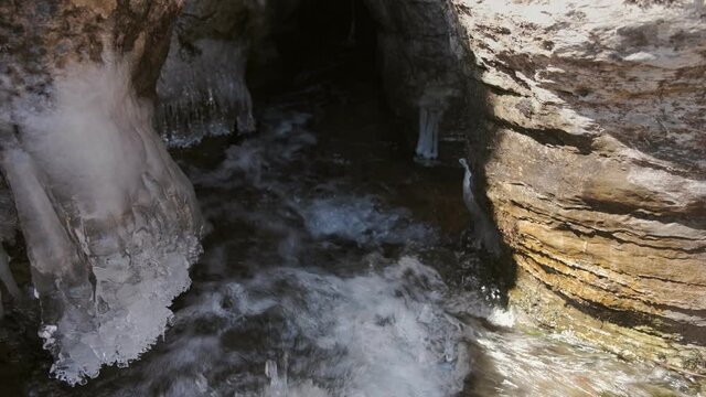 Strong mountain creek streaming in to the underground cave through the narrow crack in the textured, eroded cliff covered by icicles