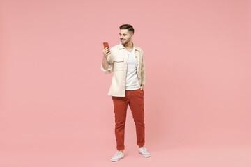 Full length young smiling happy fun trendy fashionable friendly caucasian man in jacket white t-shirt using mobile cell phone chat online in social network isolated on pastel pink background studio.