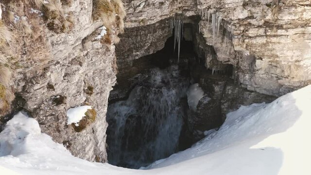 Cave in a cliff with a misterious waterfall disappearing in to the darkness of a hole with icicles hanging from the edge and snow in the foreground