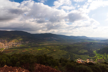 Naklejka premium A view of a mountain range and a green valley in the morning at sunrise, against a dramatic backdrop of blue skies and clouds. North District Israel. High quality photo. Travel concept hiking