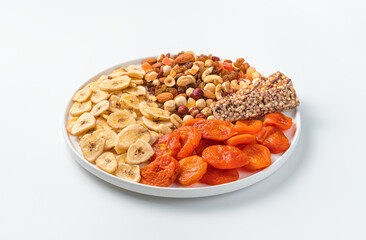 Dried apricots, dried bananas, a mixture of nuts and granola bar on a large, flat plate on a light background. Side view, close-up.