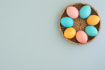 Beautiful Easter background with a wicker basket with colorful Easter eggs in pastel colors on a gentle blue background with a place for text. Top view, flat lay. Spring holiday concept