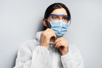 Portrait of young doctor man wearing PPE suit, putting on medical face mask against coronavirus and covid-19. Background of grey textured wall.