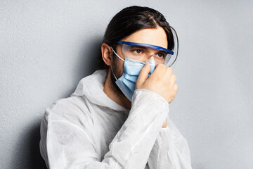 Portrait of young doctor man wearing PPE suit, putting on medical face mask against coronavirus and covid-19. Background of grey textured wall.