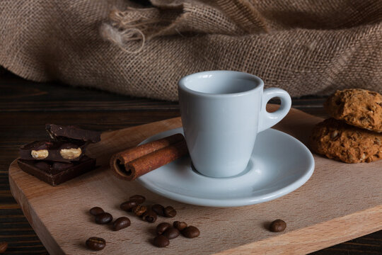 Cup of hot espresso with cinnamon stick, chocolate pieces and oatmeal cookies on wooden board, rustic burlap background. Cozy coffee break still life.