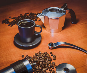 Freshly brewed cup of coffee from a moka pot, set on table in-between coffee grinder, coffee beans ant top part of moka pot.