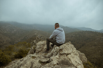 Man from behind in a gray jacket sitting on a rock on top of a mountain in autumn with mist