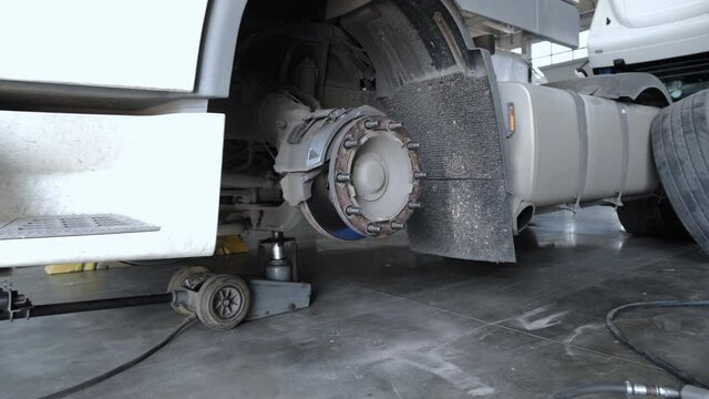 A white lorry without front wheel stands on a jack at the truck service station. Zoom out from the truck's front wheel hub. Annual maintenance