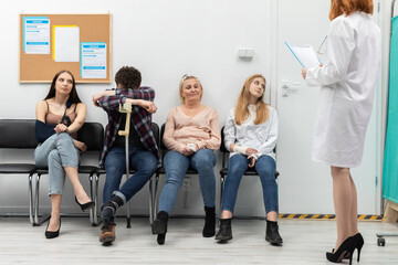 The doctor standing in front of her office reads out the next person and invites them in for a check-up. People of different age are sitting on chairs