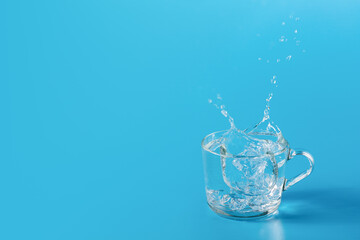 Clear cold water in a glass cup on blue background. Beautiful splash from falling ice