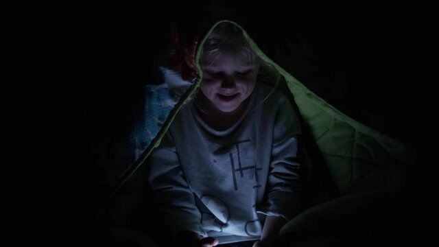 Little girl uses her cell phone at night under the blankets.