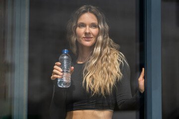 Woman in sportswear holding bottle of drinking water by the window at home. Sporty active attractive fitness woman looking outside