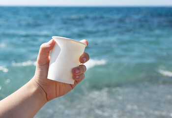 Female hand holds white cartboard coffee cup on blue sea background, takeaway coffee outside, active life and recreational concept