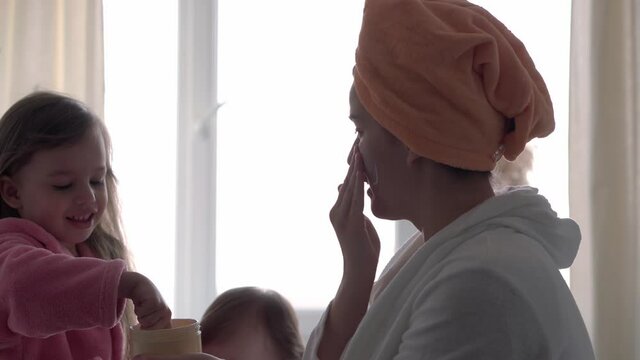 Authentic cute young woman applying face mask of baby girl. mom with 2 children daughters different ages in bathrobes after bath on white bed oposite window. Spa, motherhood, childhood, family concept