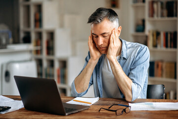 Mature caucasian gray-haired exhausted man, manager or freelancer, sitting at work desk, holding...