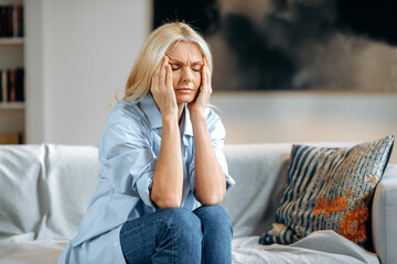 Middle aged blonde caucasian woman sits on couch at living room holding her head with her hands,...