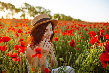 Young woman sits in the middle of the red poppy field and smells the flowers. Girl enjoys a good sunny day among the flowers. Spring and sunset concept.