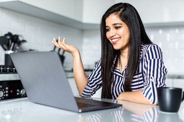 Young indian woman talking on video call and waving hand while sitting at table in the kitchen